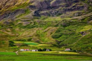 A dramatic mountain backdrop dwarfs a tiny hamlet , set amidst lush pastures, in rural Iceland.